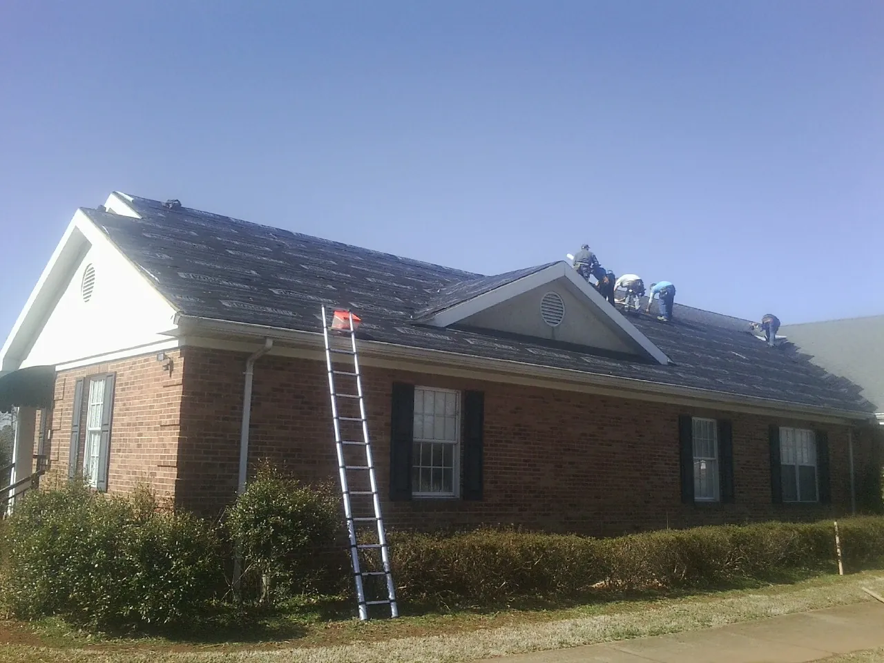 Crew tearing shingles off a brick ranch roof with a ladder set — Metal Roof Repair work by our crew in Ilion