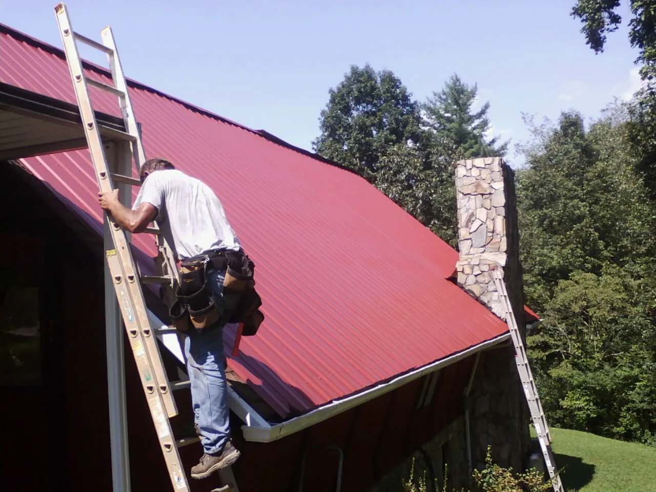 Roofer on a ladder beside a red metal roof and stone chimney — Soffit & Fascia Repair by Roofing Brothers in Ilion